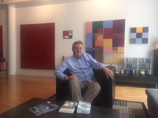 Collector Paul Gurtler seated before part of his collection. Paintings from left to right by: YM Whelan, New York artist Dulcie Dee, Blaise Delong, and Steve Rockwell